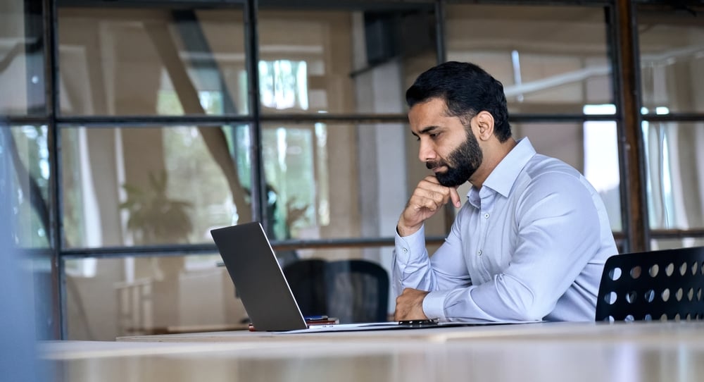 Thoughtful concentrated indian business man entrepreneur investor manager using computer, watching webinar working in office analyzing online data market thinking doing web research looking at laptop